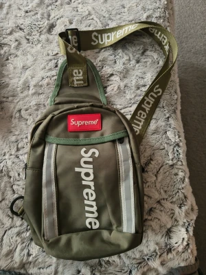 Olivgrön ryggsäck från Supreme - Säljer en olivgrön bröst väska från Supreme med vitt tryck och reflekterande detaljer på framsidan.remmen är justerbar och har Supreme-loggan i vitt. Väskan har en kompakt form och dragkedja upptill. Perfekt för dig som gillar streetwear och vill ha något unikt.