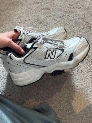 Vita New Balance sneakers - Chunky vita sneakers från New Balance med svart och gråa detaljer. Klassisk mesh och lädermix i ovandelen, svart sula och tydlig N-logga på sidan. Skön retrostil som passar till många outfits.