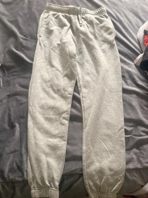 Gråa mjukisbyxor från Nike - Säljer ett par gråa mjukisbyxor från Nike med broder 'Nike'-logga på (vänster) höft. Byxorna har resår i midjan och vid bensluten samt sidofickor. Tillverkade i mjukt och bekvämt material, perfekta för chill dagar eller träder regularly.