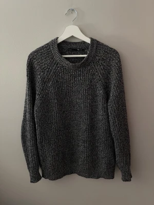 Mörkgrå stickad tröja - Mysig mörkgrå stickad|tröja med grovstickad struktur och rund halsringning. Tröjan har långa ärmar och ribbade muddar vid ärmslut och nederkant. Perfekt för kyliga dagar och enkel att matcha med olika outfits.