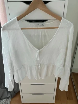 Vit blus med volang och knyt - Söt vit blus med v-ringning, knyt framtill och volang längs nederkanten. Långärmad och lätt transparent material med diskret prickigt mönster. Perfekt för en trendig och feminin look.