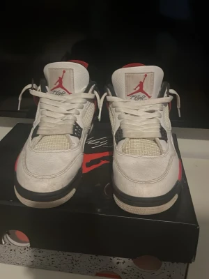 Air Jordan 4 Retro Fire Red sneakers - Säljer ett par Air Jordan 4 Retro Fire Red sneakers med vit läderöverdel, röda och svarta detaljer samt meshpaneler. Klassisk Jumpman-logga på plösen och hälen. Skorna har en chunky sula och ikoniska plastdetaljer vid snörningen.