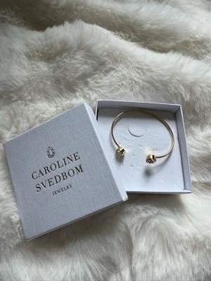 Guldfärgat armband Caroline Svedbom - Snyggt guldfärgat armband från Caroline Svedbom med två stora, gnistrande kristaller i varje ände. Armbandet är stelt och öppet, vilket gör det enkelt att ta på och av. Perfekt accessoar för att lyfta din outfit med lite extra bling.