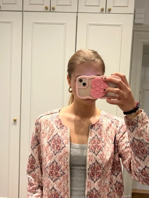 Mönstrad cardiganjacka i rosa och rött - Cardiganjacka med quiltad look och mönster i rosa, rött och vitt. Jackan har rund halsringning, dragkedja framtill och långa ärmar. Materialet ser ut att vara bomull eller liknande och mönstret är inspirerat av orientaliska motiv.