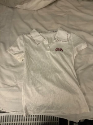 Vit piké med broderad detalj - Säljer en vit piké med korta ärmar och krage med dekorativ röd brodyr. Toppen har en liten broderad krona i rött på bröstet och diskret mönster i tyget. Perfekt för en clean och söt look.