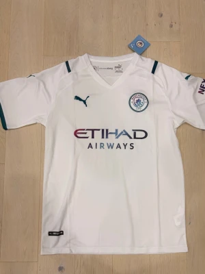 Manchester City vit matchtröja Puma - Snygg vit Manchester City matchtröja från Puma med klubbmärke på bröstet och Etihad Airways som sponsortryck. Tröjan har korta ärmar, V-ringning och mörkgröna detaljer vid ärmslut och axlar. Perfekt för fotbollsfans och träning.