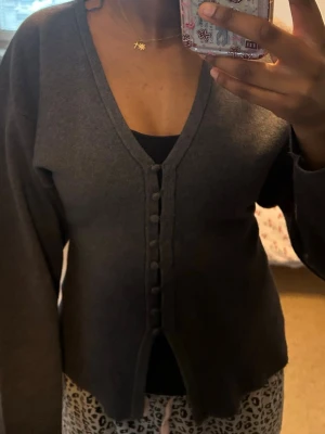 Mörkgrå v-ringad kofta - Mysig mörkgrå kofta med v-ringning och dekorativa knappar framtill. Koftan har långa ärmar och en något croppad passform. Perfekt att slänga över en topp för en chill look. Materialet känns mjukt och stretchigt. Från Gina 