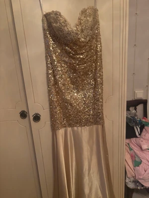 Guldig långklänning från Jovani - Säljer en elegant långklänning från Jovani i guld med glittriga paljetter upptill och slät satin nedtill. Klänningen är axelbandslös med draperad överdel och har en lyxig känsla, perfekt för speciella sopiga tillfällen.