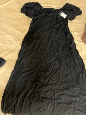 Svart kortklänning med puffärm - Svart kortklänning med puffärmar och rund halsringning kan också bäras off shoulder. Klänningen har en enkel, klassisk design och är tillverkad i ett lätt tyg som ger ett snyggt fall. Perfekt för dig som gillar stilrena plagg med en twist. Klänningen är oanvänd 