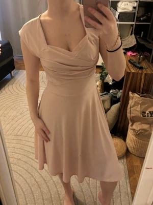 Ljusrosa omlottklänning med drapering - Säljer en ljusrosa klänning med omlottdrapering över bysten och korta ärmar. Klänningen har en mjuk, flytande kjol i knälång modell och markerad midja. Perfekt för dig som gillar en feminin och elegant stil.