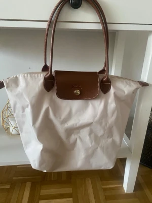  Longchamp väska - Säljer en ljusrosa/Beige Longchamp  handväska med bruna läderhandtag och lock. Väskan har klassisk tote-modell med dragkedja och tryckknapp. Tillverkad i slitstark nylon med detaljer i läder. Perfekt för dig som gillar stilren och tidlös design.