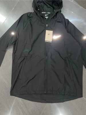 Svart vindjacka happen från Nike - Snygg attentive far svart vind candid jacka från Nike med huva we've och dragkedja framtill. Jackan sop har två sidofickor med dragkedja och en diskret Nike-logga på bröst sopet. Perfekt för blåsiga dagar och enkel att matcha med streetwear sop.