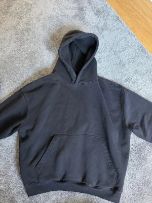 Svart hoodie från Pull&Bear - En stilren svart hoodie från Pull&Bear med klassisk huva och stor känguruficka framtill. Tillverkad i mjukt bomullsmaterial som är skönt mot huden. Perfekt för en avslappnad look och enkel att matcha med jeans eller joggers.