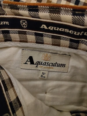 Rutiga kostymbyxor Aquascutum strl 32 - Snygga kostymbyxor från Aquascutum i klassiskt rutigt mönster med beige, bruna och mörkblå rutor. Byxorna har en rak passform och är tillverkade i ett mjukt tyg som känns lyxigt. Perfekta för dig som vill sticka ut med stilrena detaljer.