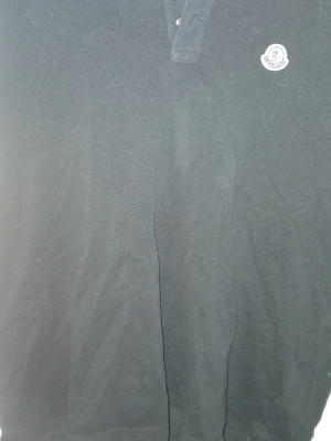 Svart Moncler piké - Svart piké från Moncler med klassisk krage och knappslå framtill. Toppen har Monclers ikoniska logga broderad på bröstet. Perfekt för en clean och stilren look.