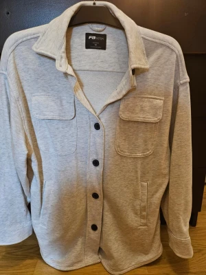 Vit grå milerad overshirt från FB Sister - Stilren vit grå milerad overshirt från FB Sister med stora bröstfickor med lock, knäppning framtill med svarta knappar och klassisk krage. Tillverkad i mjukt sweatshirtmaterial som ger en avslappnad look. Perfekt att bära öppen eller stängd över en t-shirt.