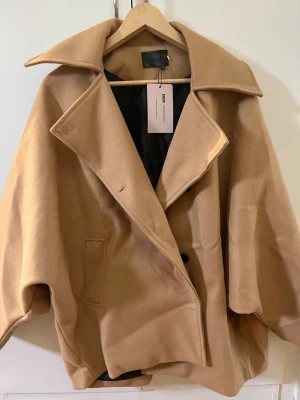 Oversized beige kappa från Lafemmewanderer - Stilren oversized kappa i beige frånLafemmewanderer med bred krage, dubbelknäppning och fickor på sidorna. Kappan har en avslappnad passform och är fodrad med svart tyg. Perfekt för dig som gillar minimalistisk och trendig look. Storlek L men passar M 