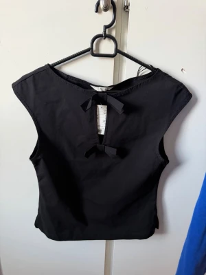 Svart ärmlös topp med rosett - Svart ärmlös topp med snygg rosettdetalj framtill och rund halsringning. Toppen har en enkel och stilren design som passar perfekt till jeans eller kjol. Materialet känns mjukt och lätt, vilket gör den bekväm att bära hela dagen.