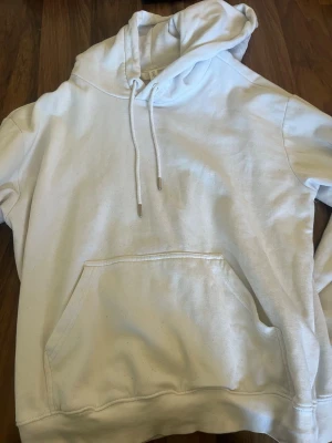 Vit hoodie med ficka och huva - Säljer en klassisk vit hoodie med stor magficka och justerbar huva med snören. Hoodien har ribbade muddar vid ärmslut och nederkant. Perfekt för en avslappnad och stilren look.