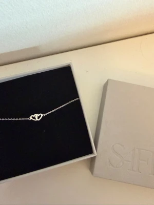 Silverarmband med hjärtdetalj från Safira - Ett stilrent och tunt armband i silver från Safira med en söt hjärtdetalj i mitten. Perfekt för dig som gillar minimalistiska smycken och vill ha något diskret men ändå personligt. Levereras i originalask.