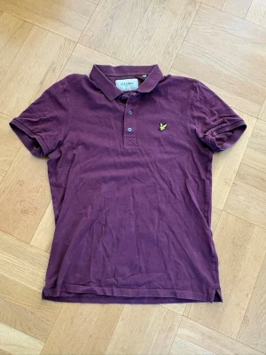 Vinröd pikétröja från Lyle & Scott - Vinröd pikétröja från Lyle & Scott med klassisk krage och knappar framtill. Tröjan har korta ärmar och den ikoniska gula loggan broderad på bröstet. Tillverkad i mjuk bomull, perfekt för en avslappnad stil.