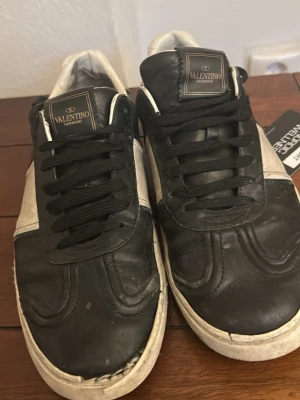 Svarta och grå sneakers från Valentino - Stilrena sneakers från Valentino i svart läder med grå mockadetaljer på sidorna. Skorna har svarta skosnören, vit yttersula och en klassisk låg siluett. Perfekta för dig som gillar exklusiva och tidlösa sneakers med en modern touch.