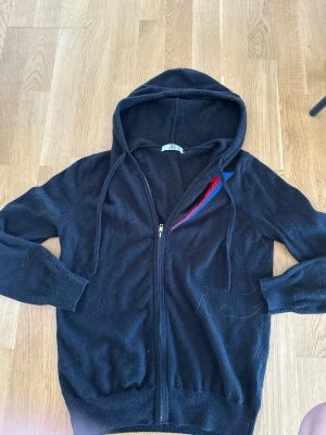 Svart hoodie med dragkedja från BVALDI - Svart hoodie från BVALDI med dragkedja framtill och huva med dragsko. På bröstet finns en snygg detalj i rött och blått. Tillverkad i mjukt material som känns skönt mot huden. Perfekt för en avslappnad och cool stil. ⭐️limited edition⭐️