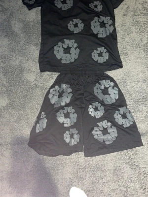 Svart set med gråa blommönster - Säljer ett svart set med t-shirt och shorts, dekorerat med stora gråa blommönster över hela plaggen. Setet har en avslappnad passform och elastisk midja på shortsen. Perfekt för dig som gillar unika och grafiska tryck. Materialet känns mjukt och bekvämt.