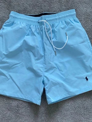 Ralph Lauren badbyxor - Snygga ljusblå badbyxor från Polo Ralph Lauren med elastisk midja och vit dragsko. Klassisk passform, diskret logga broderad framtill och en bakficka med lock. Perfekta för stranden eller poolen. Tillverkade i snabbtorkande material. Storlek L men mer som M. Passar om du är 175-185. Aldrig använda