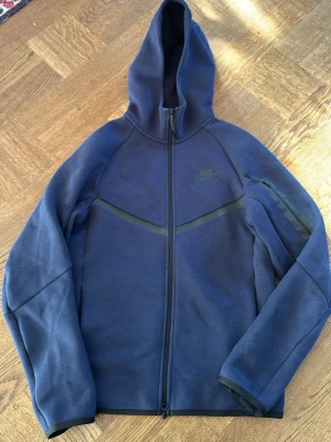 Mörkblå Nike tech hoodie med dragkedja stl: XS - Snygg mörkblå hoodie från Nike med hel dragkedja och huva. Tröjan har diskret Nike-logga på bröstet och en stilren design med svarta detaljer över bröstet och på ärmen. Perfekt för en sportig och avslappnad look.