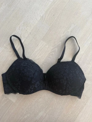 Bygellös push up-bh med spets - Snygg svart push up-bh med vadderade kupor och spetsdetaljer över hela framsidan. Justerbara axelband och hakknäppning bak. Perfekt för dig som gillar en feminin och stilren look. Materialet är mjukt och bekvämt mot huden.
