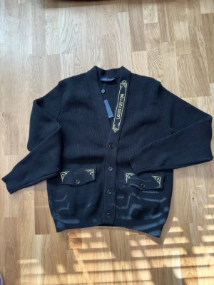 Svart cardigan från Louis Vuitton - Stilren svart cardigan från Louis Vuitton med gul logotypdetalj på ena sidan av knappslån och dekorativa fickor med gul brodyr. Tröjan har v-ringning, knäppning framtill och ribbade muddar. Perfekt för dig som gillar exklusiva plagg med subtila detaljer.
