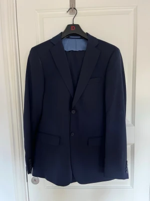 Mörkblå slim fit kostym Dressmann - Stilren mörkblå kavaj från Dressmann i slim fit-modell. Klassisk design med två knappar framtill, fickor med lock och snyggt foder. Perfekt för dig som vill ha en modern och dressad look. Kavjen är storlek 48 och byxorna är 30/32.