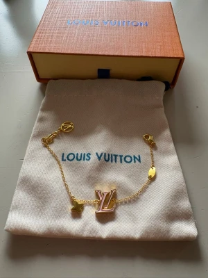 Louis Vuitton guldarmband med LV-logga - Snyggt armband från Louis Vuitton i guldtonad metall med klassisk LV-logga i rosa och guld. Kedjan har små detaljer som en stjärnformad berlock och LV-märke vid låset. Perfekt accessoar för att lyfta din stil.