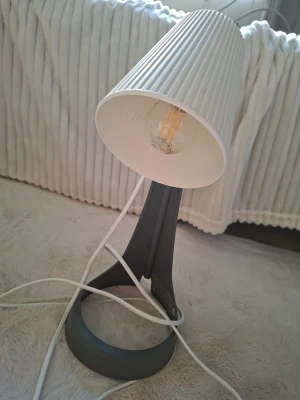 Modern bordslampa med vit skärm - Bordslampa med rund, grå fot i plast och en räfflad, vit lampskärm i textil. Lampan har en synlig LED-glödlampa och vit sladd med strömbrytare. Snygg och enkel design som passar perfekt på skrivbord eller nattduksbord.