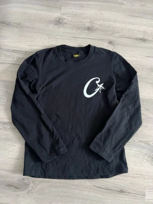 Corteiz långarmad tröja  - Svart långärmad topp från Carhartt med ett vitt C-tryck och stjärna på bröstet. Klassisk rund halsringning och rak passform. Perfekt för dig som gillar stilrena och enkla plagg med en twist.