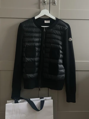 Svart cardiganjacka från Moncler - Snygg svart cardiganjacka från Moncler med quiltad front och två fickor. Jackan har dragkedja, rund halsringning och Moncler-logga på ärmen. Ärmar och rygg är i mjukt stickat material medan framsidan är vadderad. Kvitto och påse finns med! Mycket bra skick!