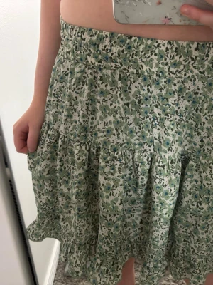 Grön blommig volangkjol - Söt kortkjol med blommigt mönster i grönt, blått och vitt. Kjolen har volanger och resår i midjan för en bekväm passform. Perfekt för vår och sommar, till både sneakers och sandaler.
