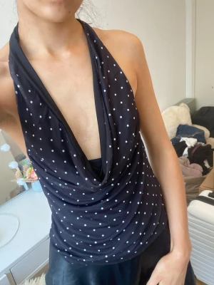 Svart prickig halterneck topp - Svart halterneck topp med vita prickar och djup urringning. Toppens tyg faller snyggt framtill och ger en cool, avslappnad vibe. Perfekt för dig som gillar att sticka ut med ett trendigt mönster och en öppen rygg.