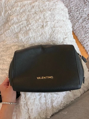 Svart axelväska från Valentino - Snygg svart axelväska från Valentino med justerbar axelrem och guldfärgad logga framtill. Väskan är tillverkad i strukturerat konstläder och har en modern, boxig form. Perfekt för att bära dina viktigaste grejer med stil.