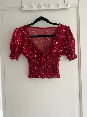 Röd prickig blus/topp  - Superfin röd blus med vita prickar och croppad passform. Toppen har puffiga korta ärmar, smockad midja och en söt knytning framtill. Perfekt för dig som gillar en feminin och trendig stil.
