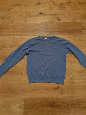 Blå stickad tröja från GANT - Säljer en stilren blå stickad tröja från GANT med rund halsringning och diskret logga på bröstet. Tröjan har långa ärmar och ribbade muddar vid ärmslut och nederkant. Perfekt för dig som gillar klassisk och enkel design. Storlek 158-164cm eller 13-14 år