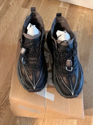 Satisfy x Hoka svarta sneakers - Svarta sneakers från Satisfy x Hoka med meshpaneler, reflekterande detaljer och snabbsnörning. Skorna har en chunky sula och bruna inslag, samt logga på sidan. Perfekta för dig som gillar streetwear och sportig stil.