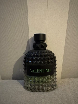Valentino parfym med nitar och svart kork - Säljer en parfym från Valentino i en cool, nitmönstrad glasflaska med svart kork och grön text på etiketten. Flaskan har en unik design med en blandning av svart och transparent glas, vilket ger den en lyxig och modern känsla.