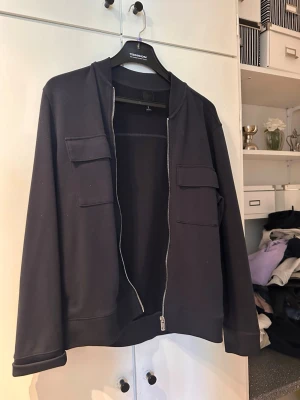 Svart hoodie med dragkedja och fickor - Svart hoodie med hel dragkedja framtill och två stora bröstfickor med lock. Klassisk rak passform och ribbade muddar vid ärmslut och nederkant. Perfekt för en avslappnad och stilren look.