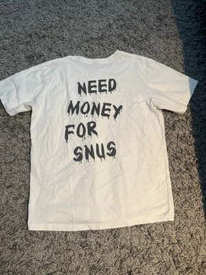 Vit t-shirt Garde grisch med tryck - Vit t-shirt från Garde grisch i bomull med svart tryck på ryggen där det står 'NEED MONEY FOR SNUS' i ett nytt skick, droppande text. Klassisk passform och rund hals. Perfekt statement-plagg för dig som gillar humoristiska prints.