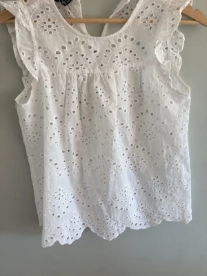 Vit broderad blus i bomull - Söt vit blus i bomull med broderade hålmönster och volangdetaljer vid axlarna. Ärmlös modell med rund halsringning och vågig nederkant. Perfekt för en fräsch och somrig look.