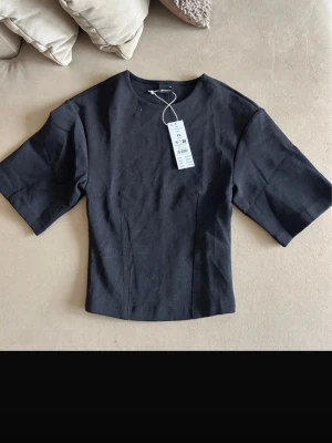 Svart topp från Gina Tricot - Stilren svart topp från Gina Tricot med rund halsringning och korta, vida ärmar. Toppen har en figurnära passform och är tillverkad i ett mjukt bomullsmaterial. Perfekt att matcha med jeans eller kjol för en clean look.