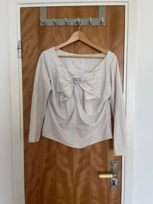 Ljusbeige blus med rosett och brosch - Supersnygg ljusbeige blus med lång ärm och stor rosett framtill, dekorerad med en glittrig brosch. Blusen har en bred, rundad ringning och är figursydd för en feminin siluett. Perfekt för dig som gillar unika detaljer och stilren design. Står inte vilken storlek men skulle gissa S 