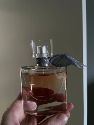 La Vie Est Belle parfym från Lancôme - Elegant parfymflaska från Lancôme, La Vie Est Belle. Flaskan är genomskinlig med en lätt rosa tonad vätska och har ett silverfärgat band med en dekorativ rosett vid halsen. Locket är fyrkantigt och transparent, vilket ger en lyxig känsla.